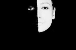 Black and white of weird enigmatic female with painted face matching color of background looking at camera in modern studio